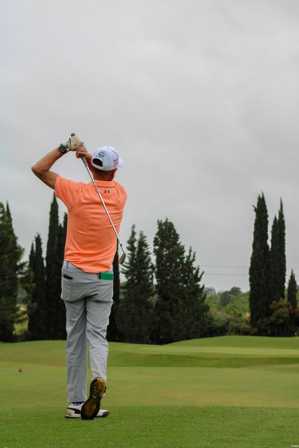 Hombre de espaldas jugando golf, con remera naranja y gorra blanca, en plena salida desde la calle de un campo de golf con cipreses de fondo.