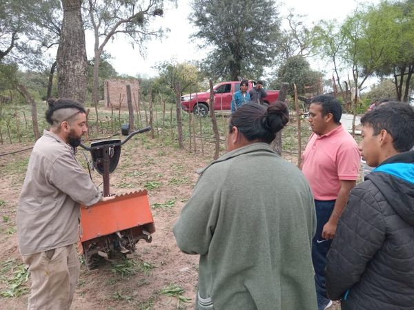 Hombre con motocultor naranja explica su funcionamiento a un grupo de pobladores en un campo con cercos de postes y un camión rojo al fondo.