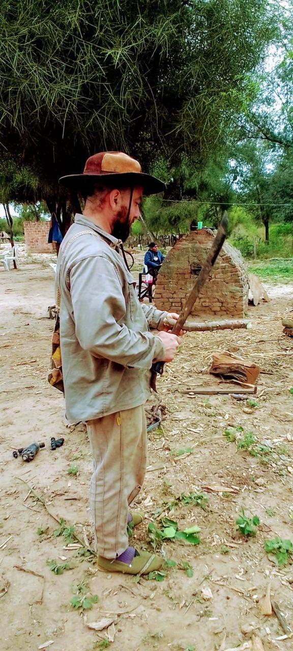 Hombre con sombrero y barba trabaja con una herramienta de mango largo junto a un horno de adobe, en un campo con árboles y tierra.