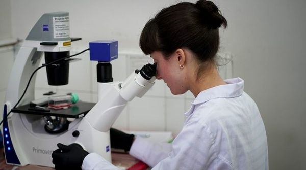 Científica con guardapolvo blanco y guantes observa una muestra en un microscopio de laboratorio