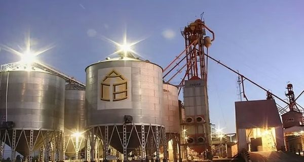 Silos metálicos de acopio con un logo en forma de casa sobre uno de ellos, y una torre de secado con caños y equipos rojos al costado