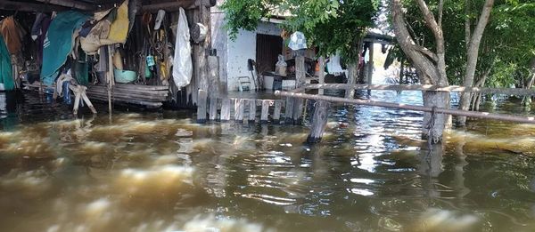 Casa rural inundada con agua turbia hasta la base de los postes y corrales de madera, con ropa colgada y trastos bajo techo de chapa