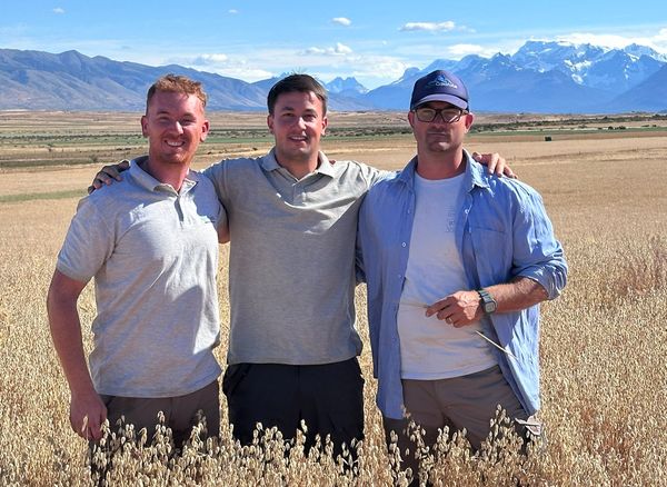 Tres hombres posan sonrientes abrazados en un campo de cultivo seco y dorado, con montañas nevadas de fondo.
