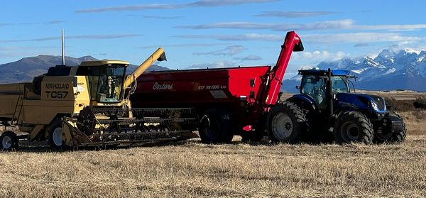 Cosechadora New Holland TC57 descargando granos en una tolva roja Costa junto a un tractor azul New Holland en un campo de rastrojo