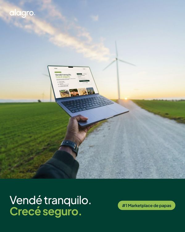 Una mano sostiene una notebook con la web de alagro, un marketplace de papas, con un camino rural, campos verdes y aerogeneradores de fondo.