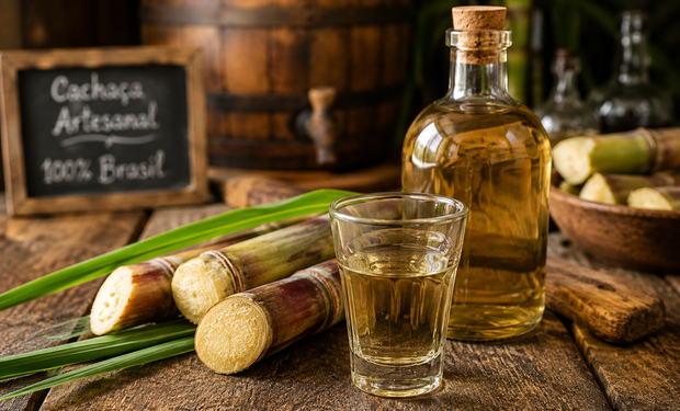 Garrafa e copo de cachaça sobre mesa rústica ao lado de cana-de-açúcar, representando a produção brasileira da bebida.