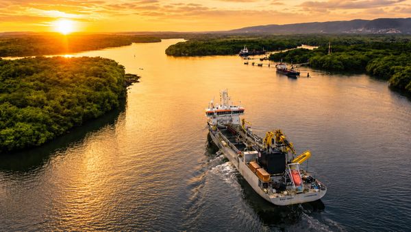 Draga de succión con brazos amarillos navegando por un río ancho flanqueado de islas y vegetación, con otras embarcaciones al fondo