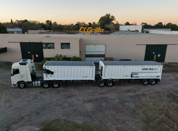 Bitren blanco con dos acoplados tolva de descarga, uno con el logo de un toro, estacionado frente a una nave industrial; se lee "Bitren Tolva 60 m³