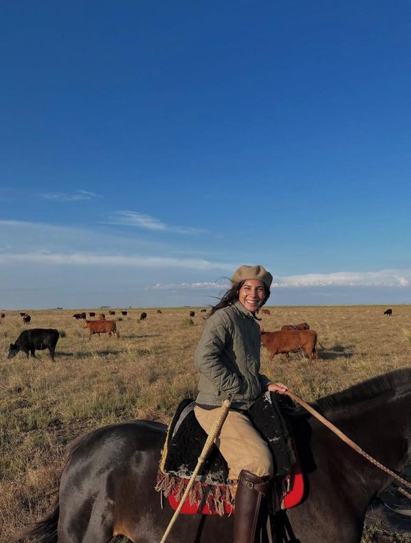 Mujer sonriente montada a caballo en un campo con vacunos, rumbo a la nota sobre el cambio de vida al campo