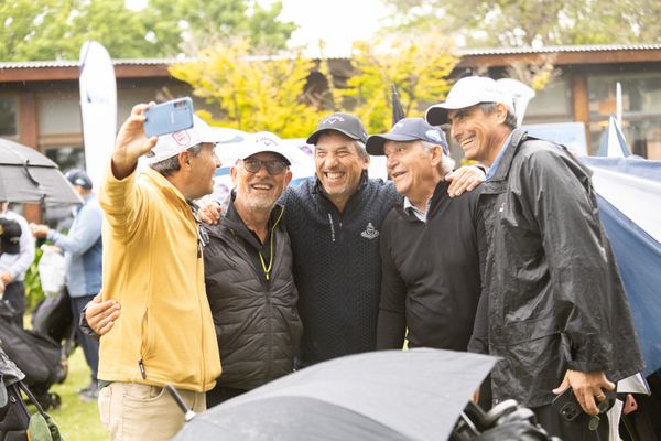Cinco hombres posan sonrientes para una selfie en un evento de golf, algunos con gorras y paraguas bajo la lluvia