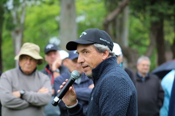 Hombre con gorra y campera de punto habla con micrófono ante un grupo de personas en un torneo de golf