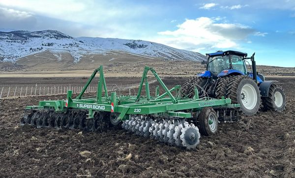 Tractor azul con una rastra de discos verde "Super Bong" trabajando un lote arado en zona de cerros nevados