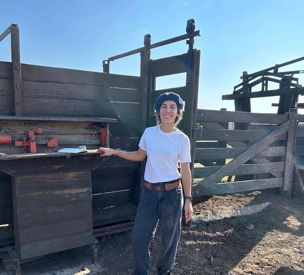 Mujer joven con gorra azul y remera blanca junto a un corral de madera con balanza y manga de manejo de hacienda.