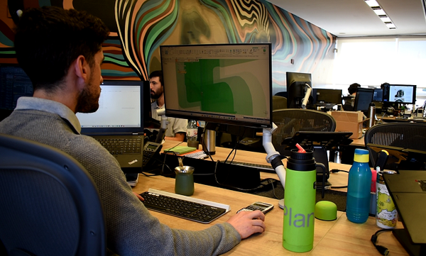 Hombre frente a una computadora en una oficina de tecnología, con un termo verde y otros objetos sobre el escritorio.