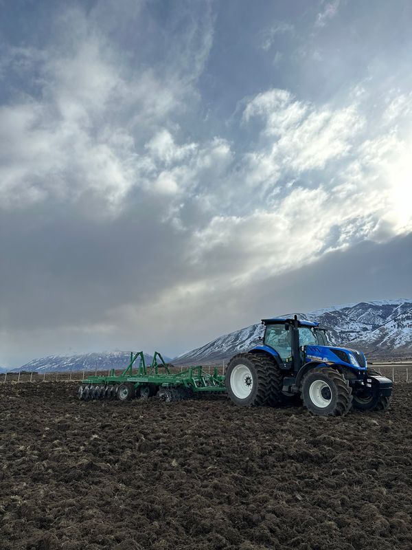 Tractor azul con rastra de discos verde trabajando un lote arado, con cordón montañoso nevado al fondo.