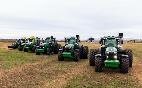 Fila de tractores verdes con ruedas dobles estacionados en un campo, con exhibición de maquinaria de fondo